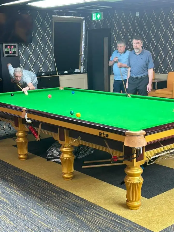 A player cueing while two others watch from the end of the table, scoreboard on the wall