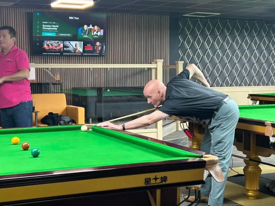 A player reaching across the table for a shot, with snooker on the screen behind